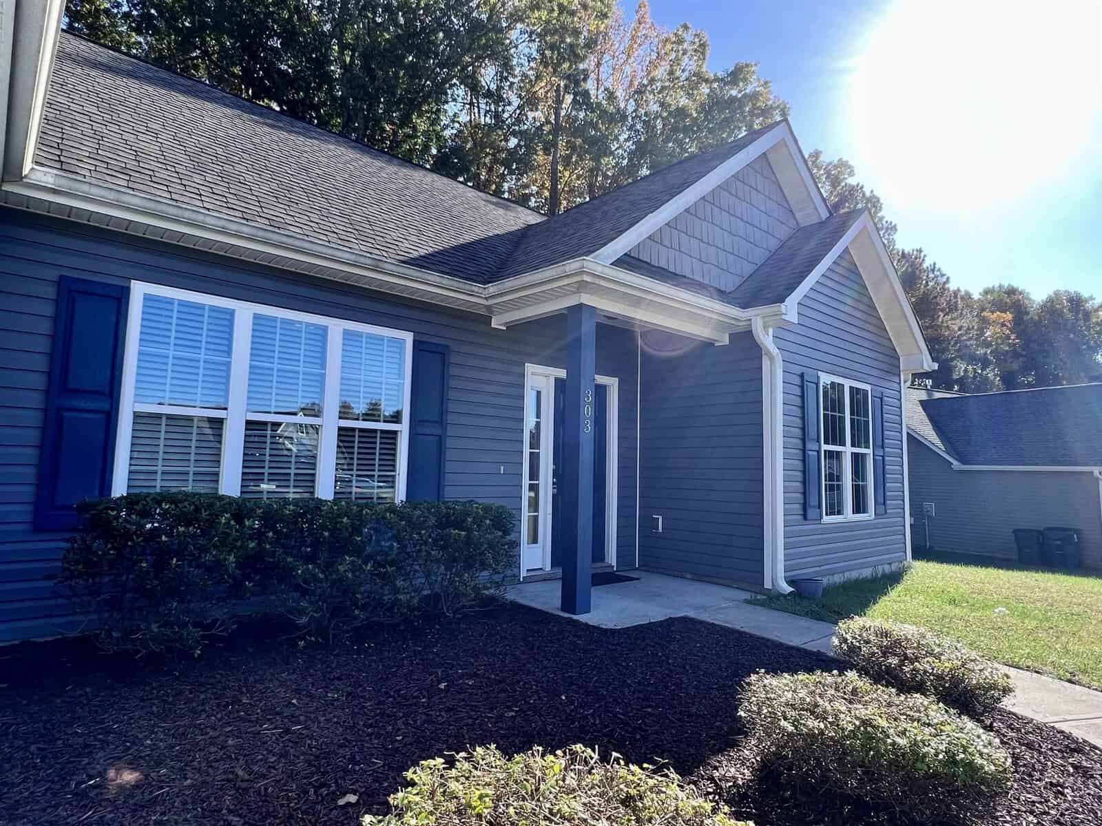 A small, single-story house with blue siding and white trim, featuring a gabled roof and a front porch. There are shrubs in front, and the sun is shining brightly in the clear blue sky.
