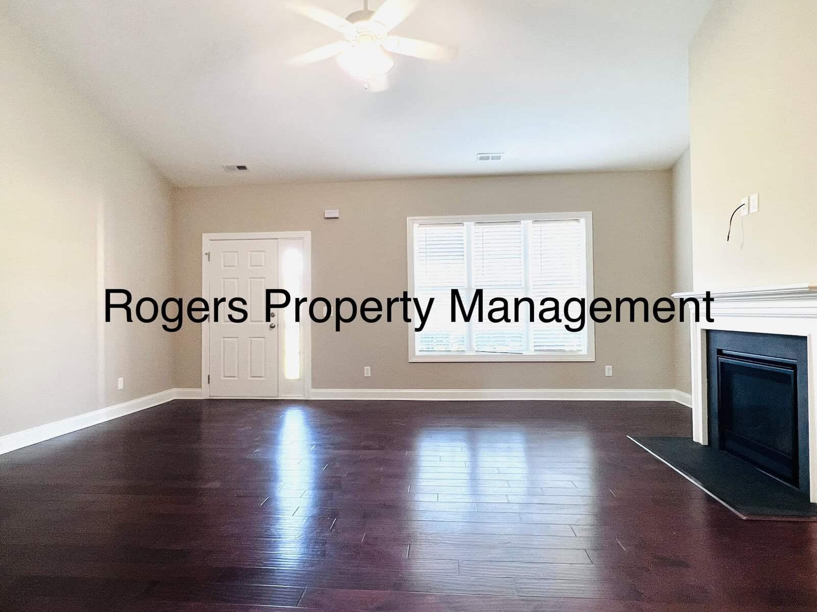 An empty living room with wooden flooring, a ceiling fan, and a built-in fireplace on the right. Large windows with blinds let in natural light. Text reads "Rogers Property Management.
