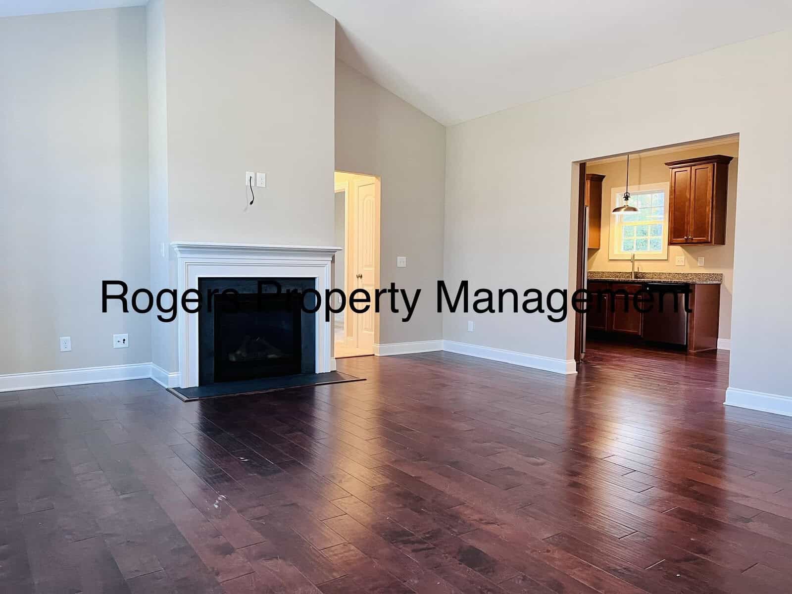 A spacious living room with dark hardwood floors and a white fireplace. The room opens into a kitchen with wooden cabinets. Text overlay reads "Rogers Property Management." The walls are painted light beige, and there's a doorway leading to another room.