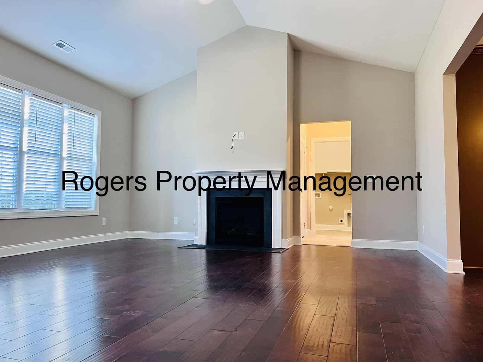 A spacious living room with dark hardwood floors and light-colored walls. A fireplace is centered in the room beneath a vaulted ceiling. Sunlight streams through a set of large windows on the left. The words "Rogers Property Management" are overlaid.