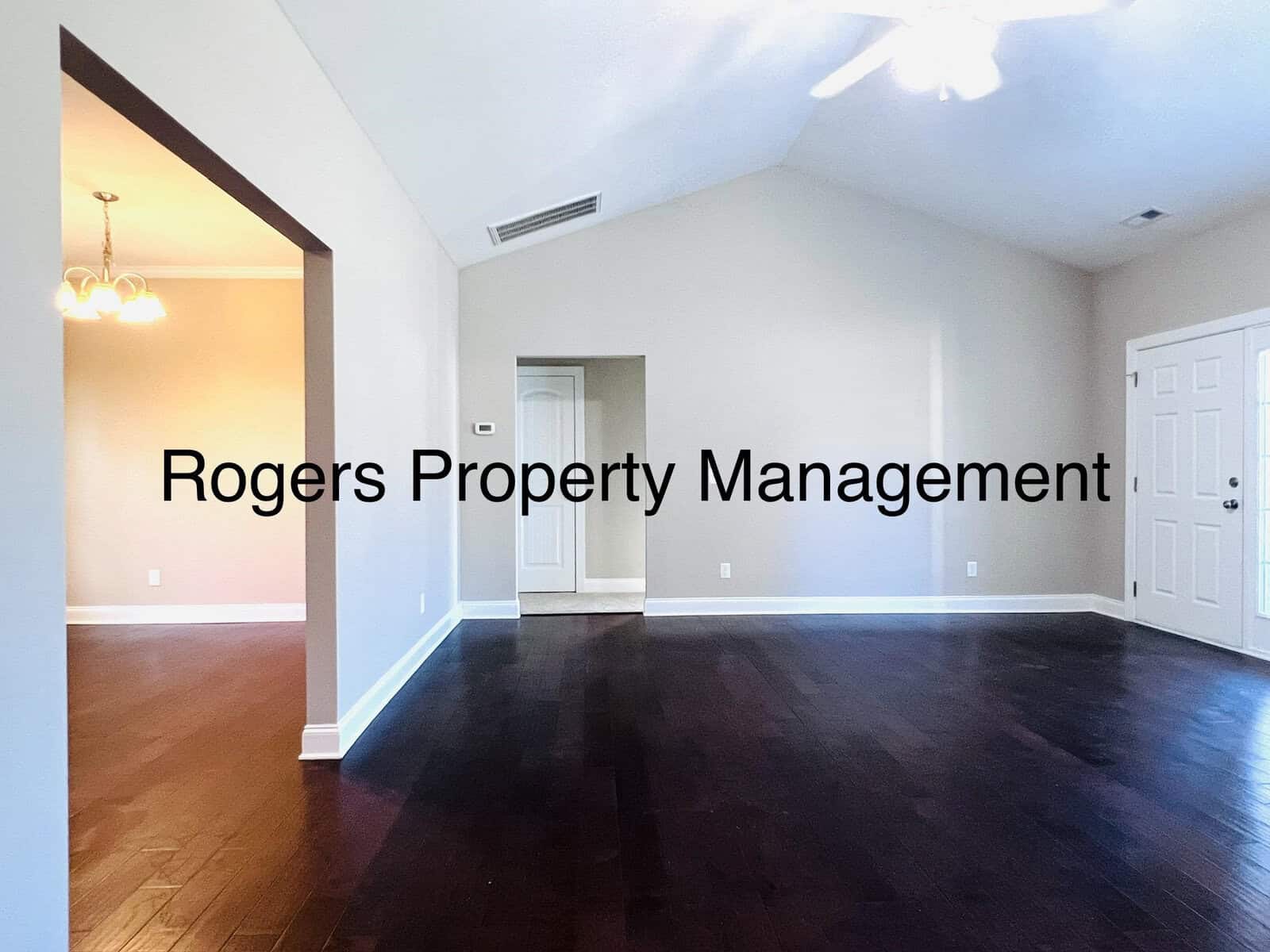 Interior of a house with dark hardwood floors, beige walls, and a high ceiling. There's a doorway to another room on the left, a closed white door on the right, and recessed lighting. Text on the image reads "Rogers Property Management.