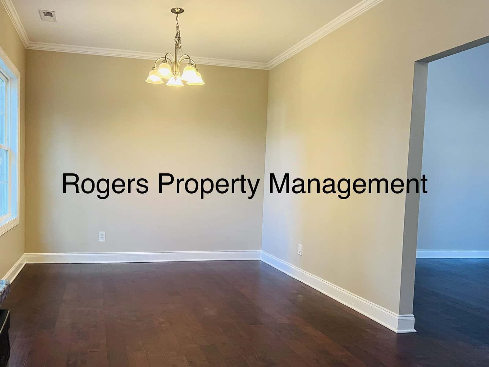 A vacant room with cream-colored walls, dark wood flooring, and a hanging light fixture. The room is well-lit by a window on the left. The text "Rogers Property Management" is overlaid in the center.