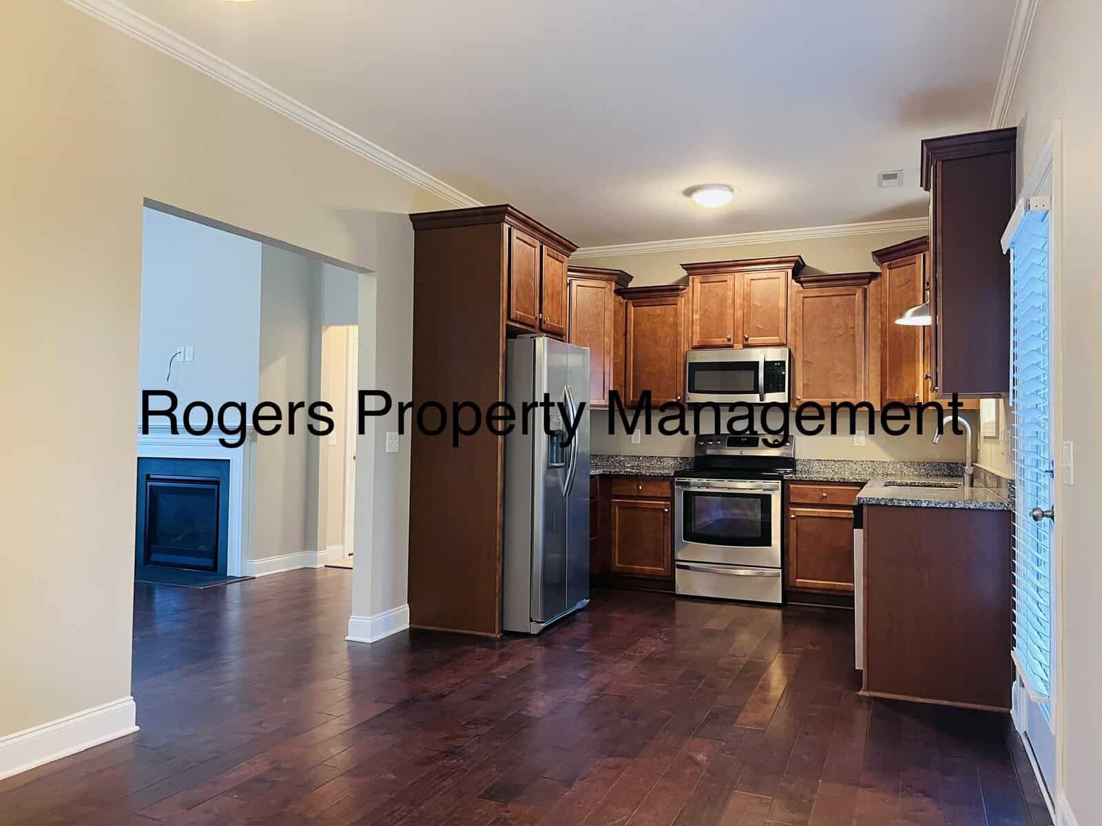 A modern kitchen with dark wood cabinets, stainless steel appliances, and granite countertops. The kitchen opens into a living area with a fireplace. Text overlay reads, "Rogers Property Management.