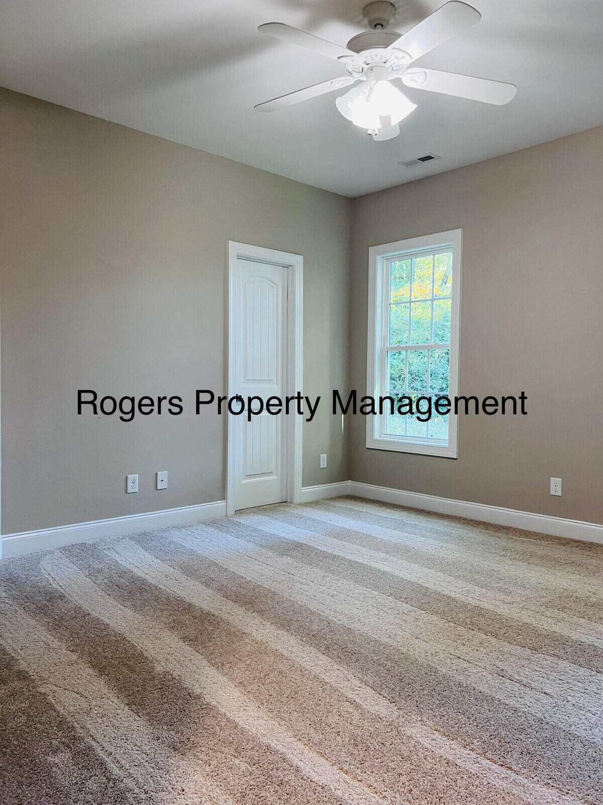 Empty room with beige walls and striped carpet. It has a white ceiling fan and a window letting in natural light. A closed door is visible on one wall. Text on the image reads "Rogers Property Management.