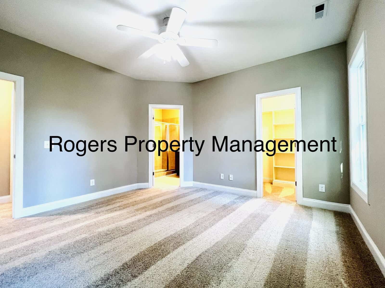 Empty room with striped carpet, beige walls, ceiling fan, and three open doorways leading to lit closets. Large window on the right wall. Text reads "Rogers Property Management.