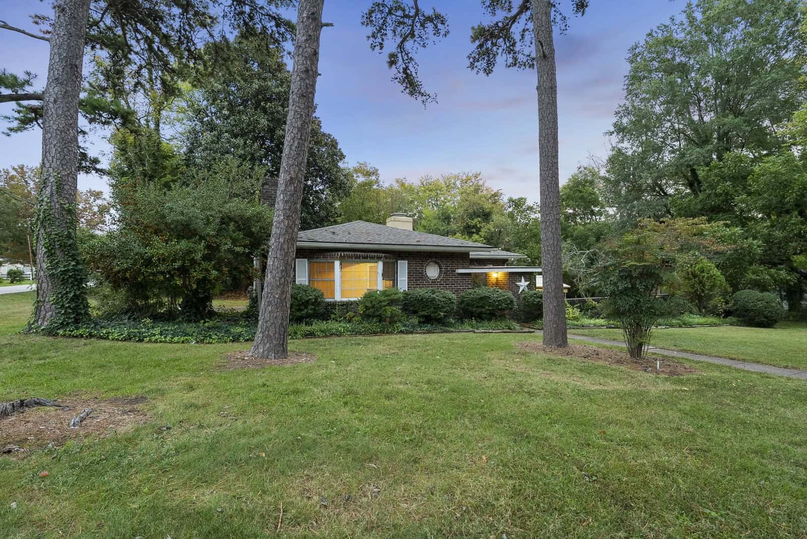 A single-story house at 346 Willowood Drive, Henderson NC, is partially obscured by trees with illuminated windows at dusk.