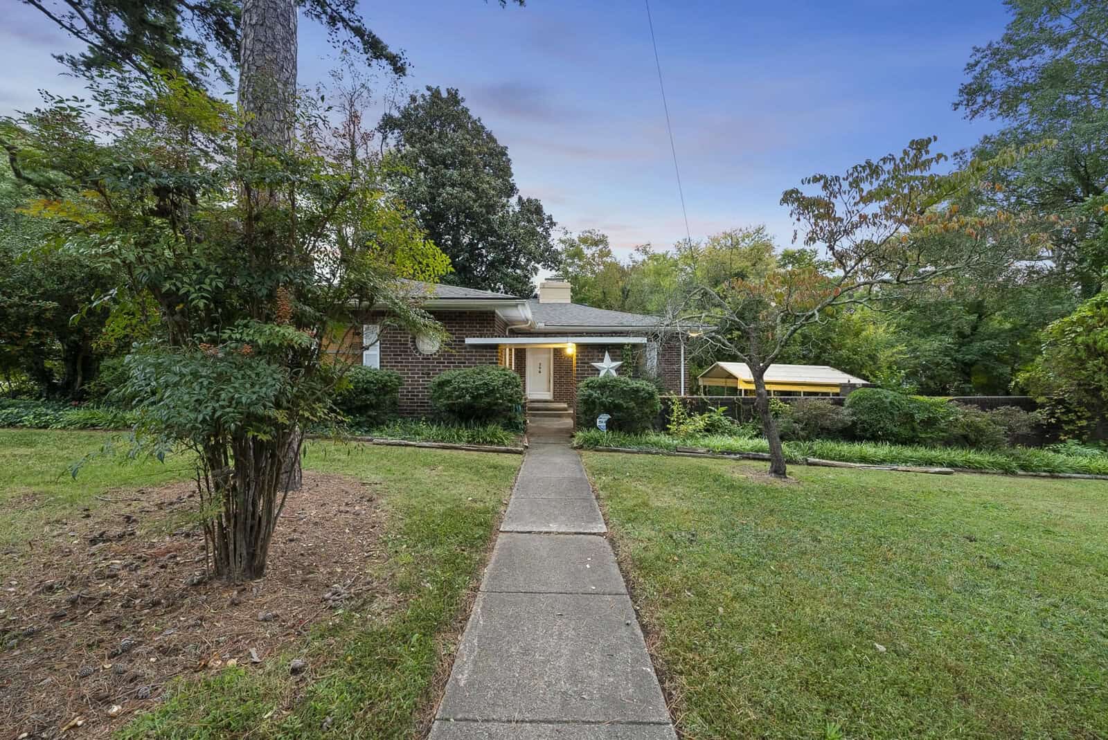 A single-story suburban home with a brick facade and white trim, featuring a central walkway leading to the front door, flanked by a lawn and various shrubs, taken at dusk.