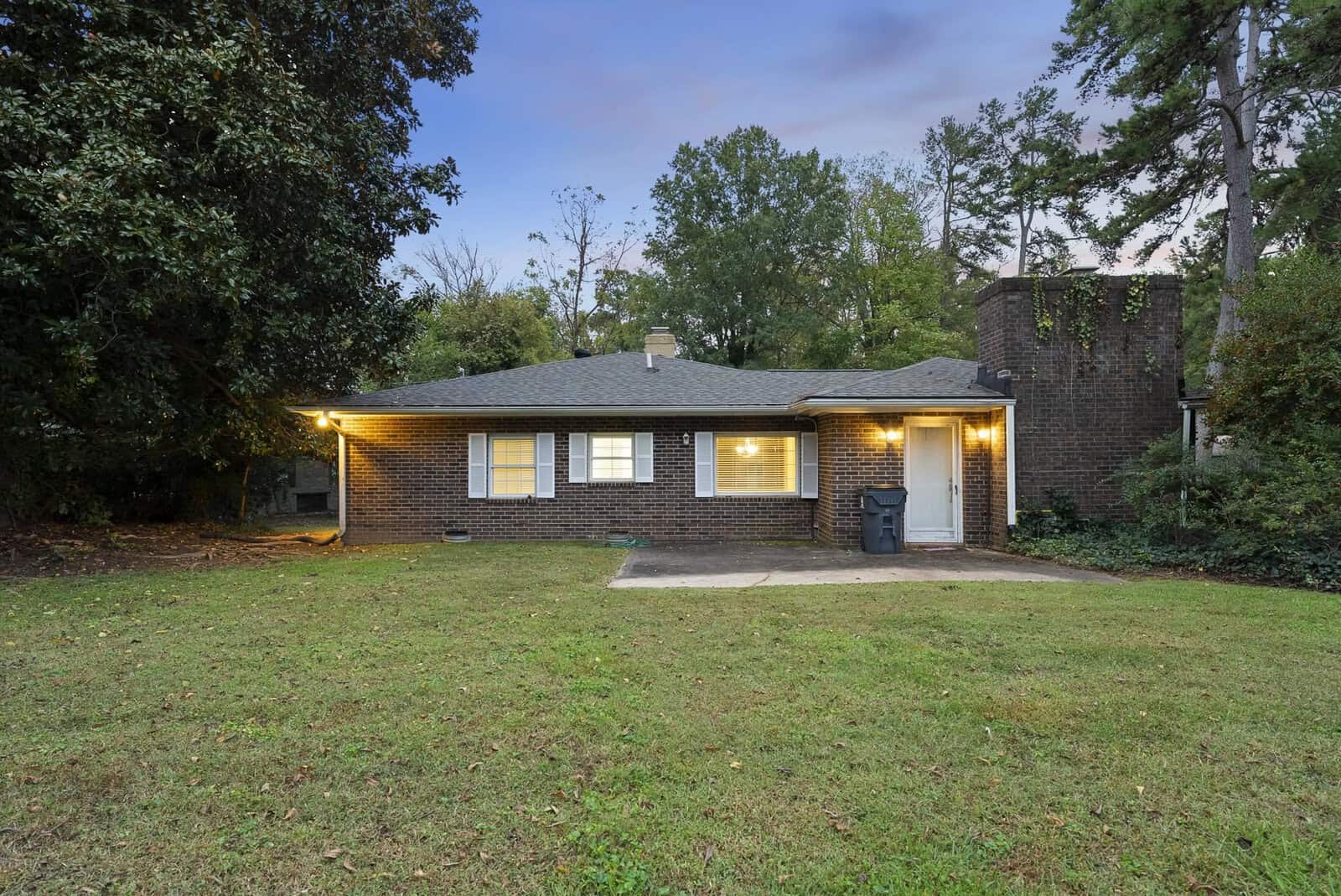 A single-story brick house with lit exterior lights, featuring a prominent chimney and a driveway without a car, set against a background of trees and a dusk sky.