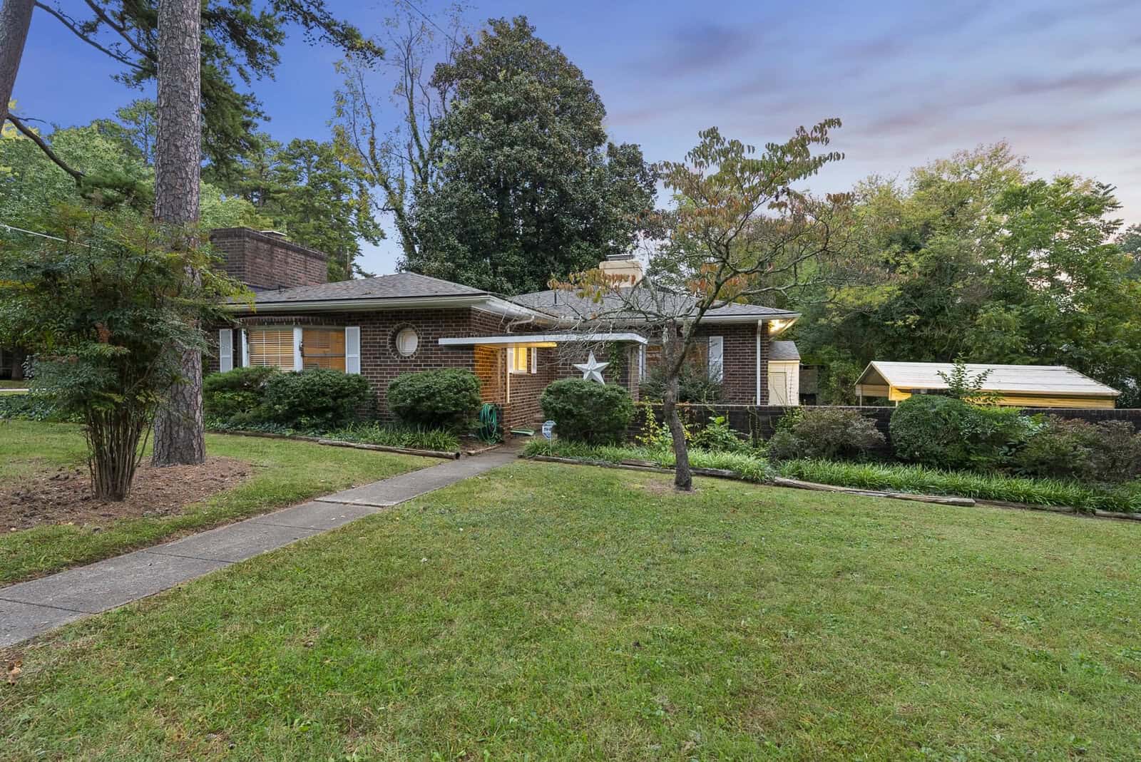 A single-story brick residential house with white accents, a well-maintained lawn, and a walkway leading to the front door, photographed during twilight with a pastel-colored sky in the background.