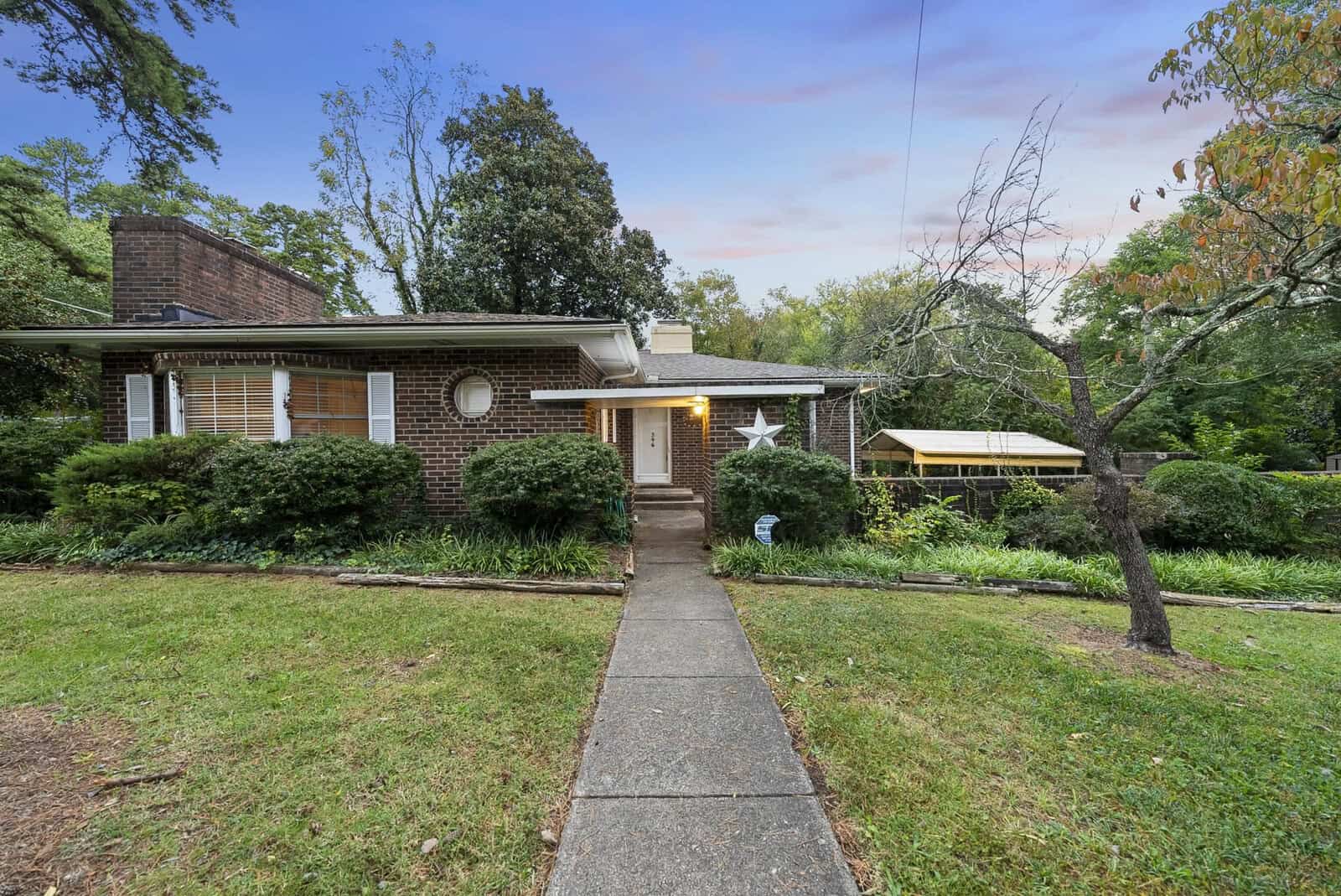 A single-story brick home with white trim, featuring a small porch area and a pathway leading to the front door, surrounded by a front yard with greenery and trees.