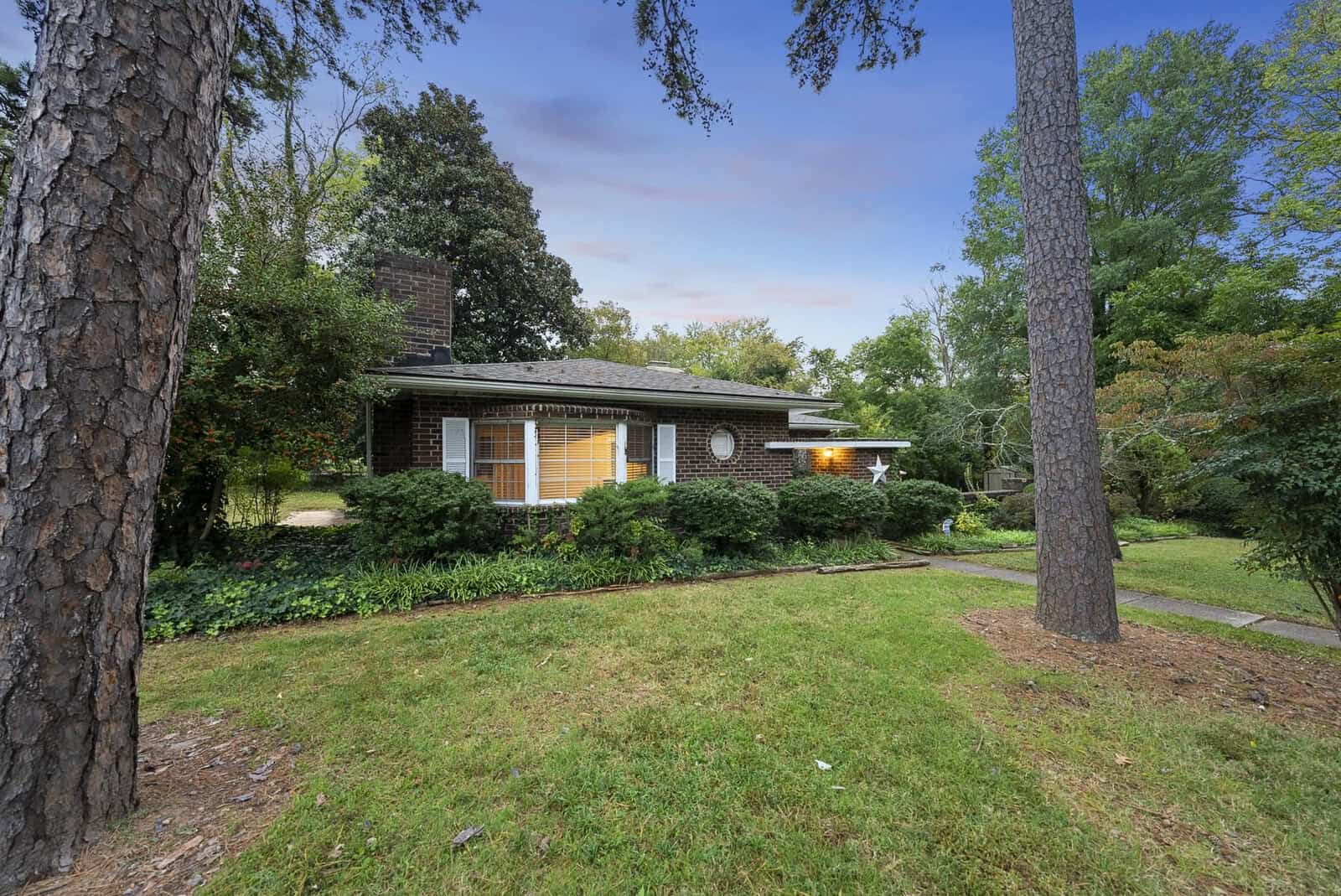 A single-story brick house with a dark shingle roof, surrounded by mature trees and greenery, under a partly cloudy sky.