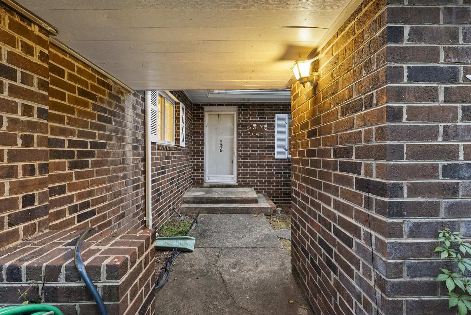 A narrow brick-walled passage leading to a white door of a residential building, illuminated by an overhead light fixture.