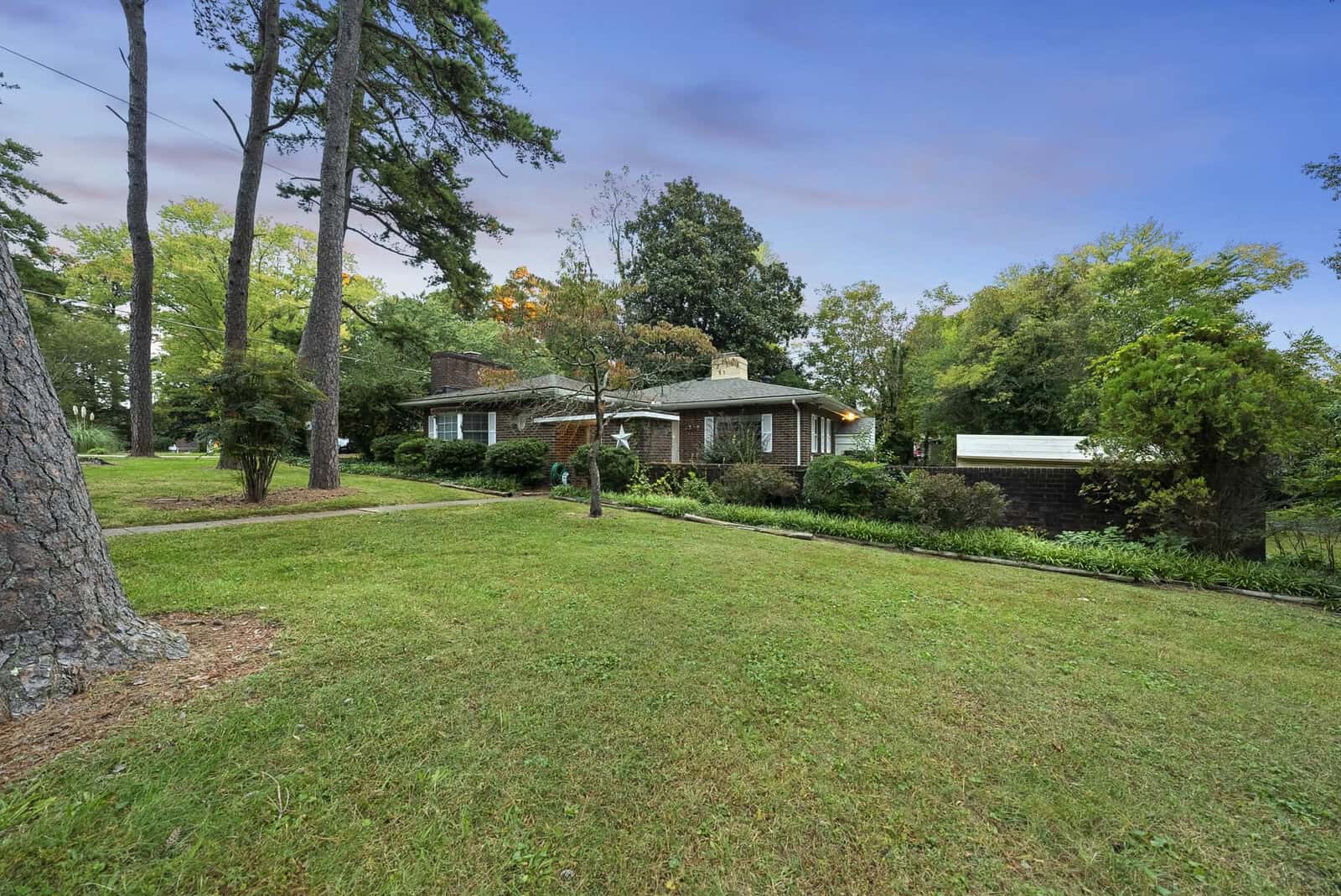 A single-story brick house with a shingled roof, surrounded by a well-manicured lawn and mature trees, under a partly cloudy sky.