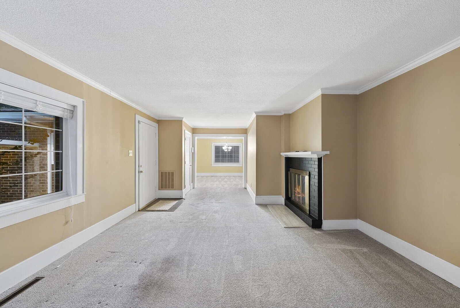 A spacious, empty room featuring beige walls, a gray carpeted floor, a fireplace on the right side, windows allowing natural light, and multiple doorways leading to adjoining rooms.