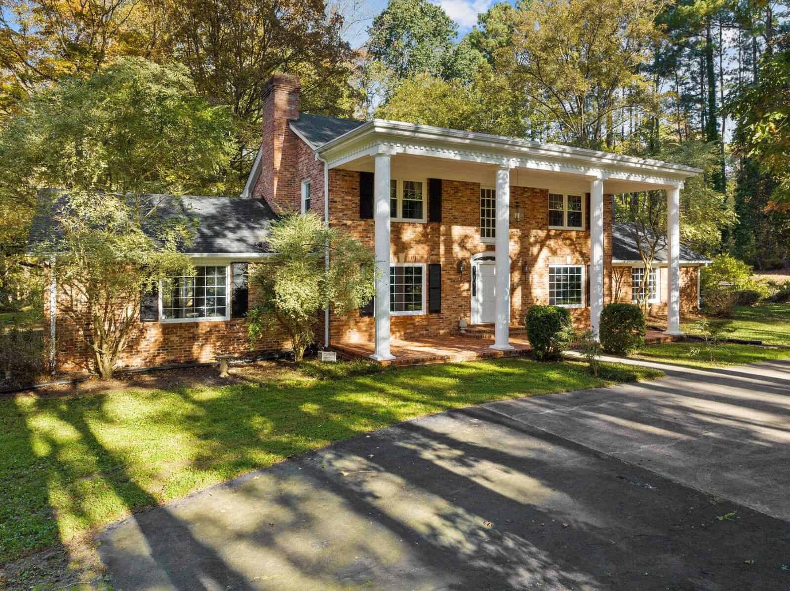 A two-story brick colonial house at 227 Salem Road features tall white columns, surrounded by the lush greenery of Oxford. The driveway gracefully curves in front of the entrance, where sunlight filters through the branches, casting gentle shadows on the lawn and pavement.