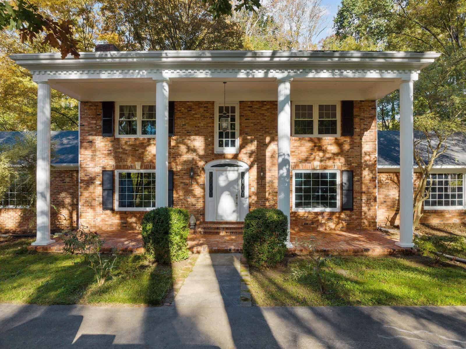 A two-story brick house with large white columns at the front entrance. It has black shutters and a white door. The surrounding area features green grass and trees with autumn leaves, casting shadows on the building.