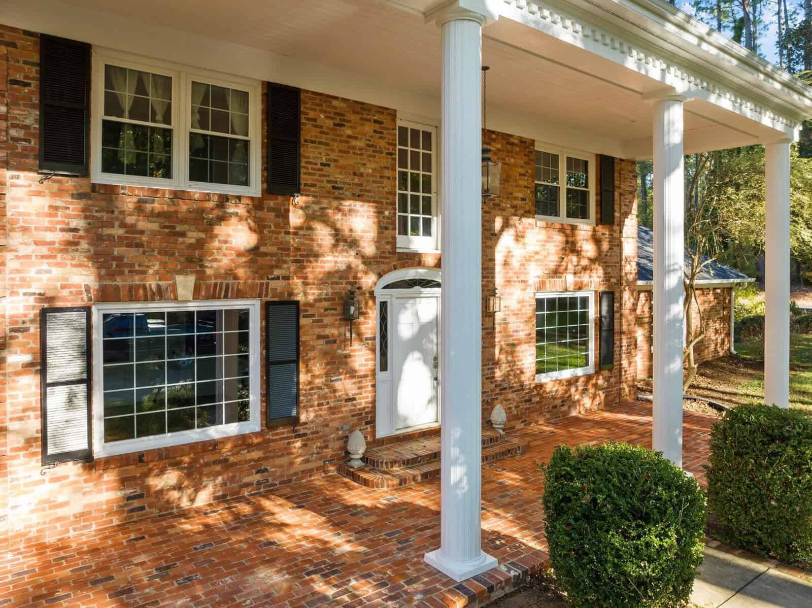 A brick house with a grand white columned portico and large windows. The building is surrounded by foliage, and sunlight casts shadows on the front facade. A neatly trimmed bush sits in front of the entrance.
