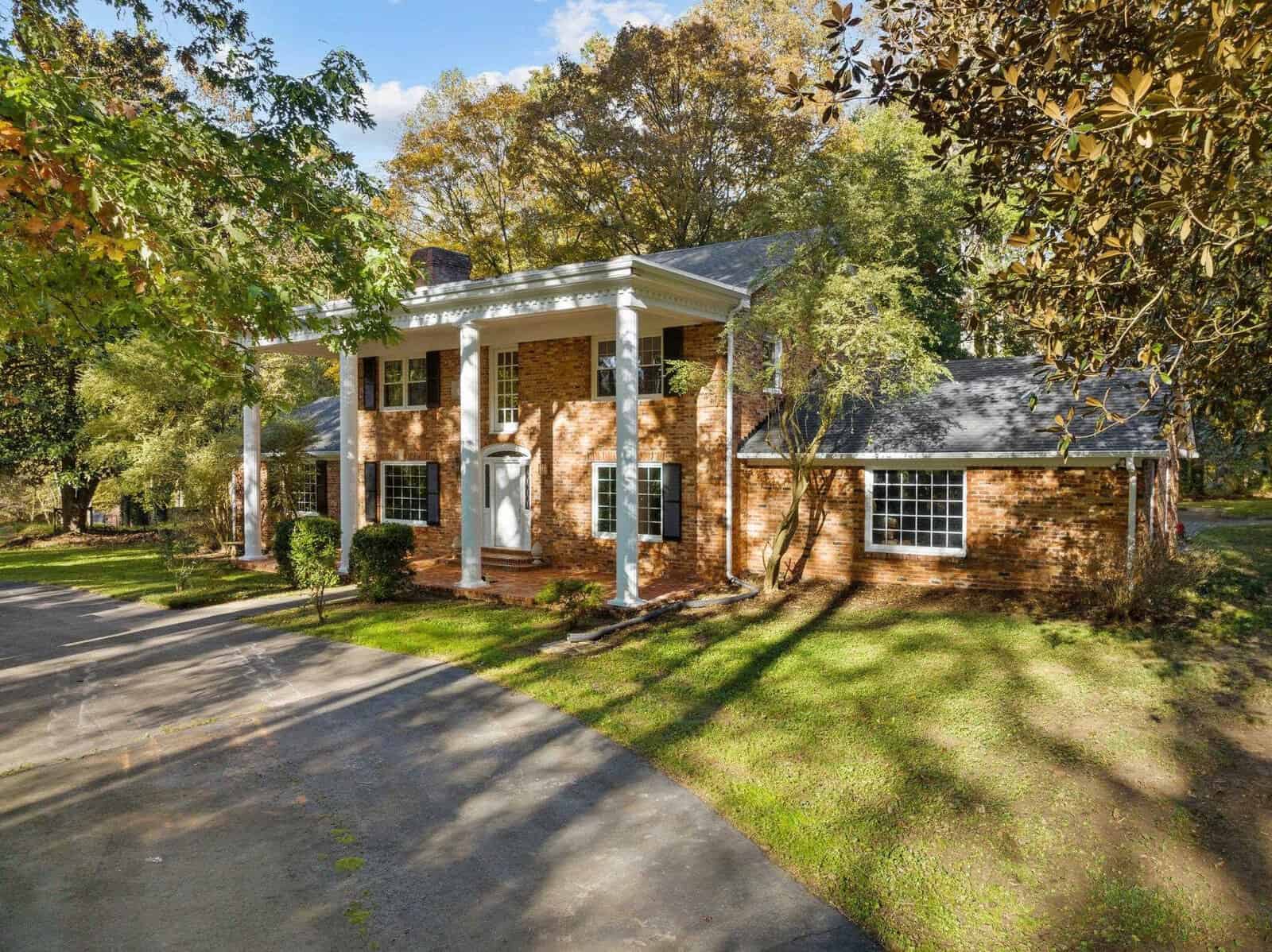 A two-story brick house with white columns, surrounded by trees and a green lawn. A curved driveway leads to the front entrance. The sky is partly cloudy, and the scene is sunlit, suggesting a pleasant day.