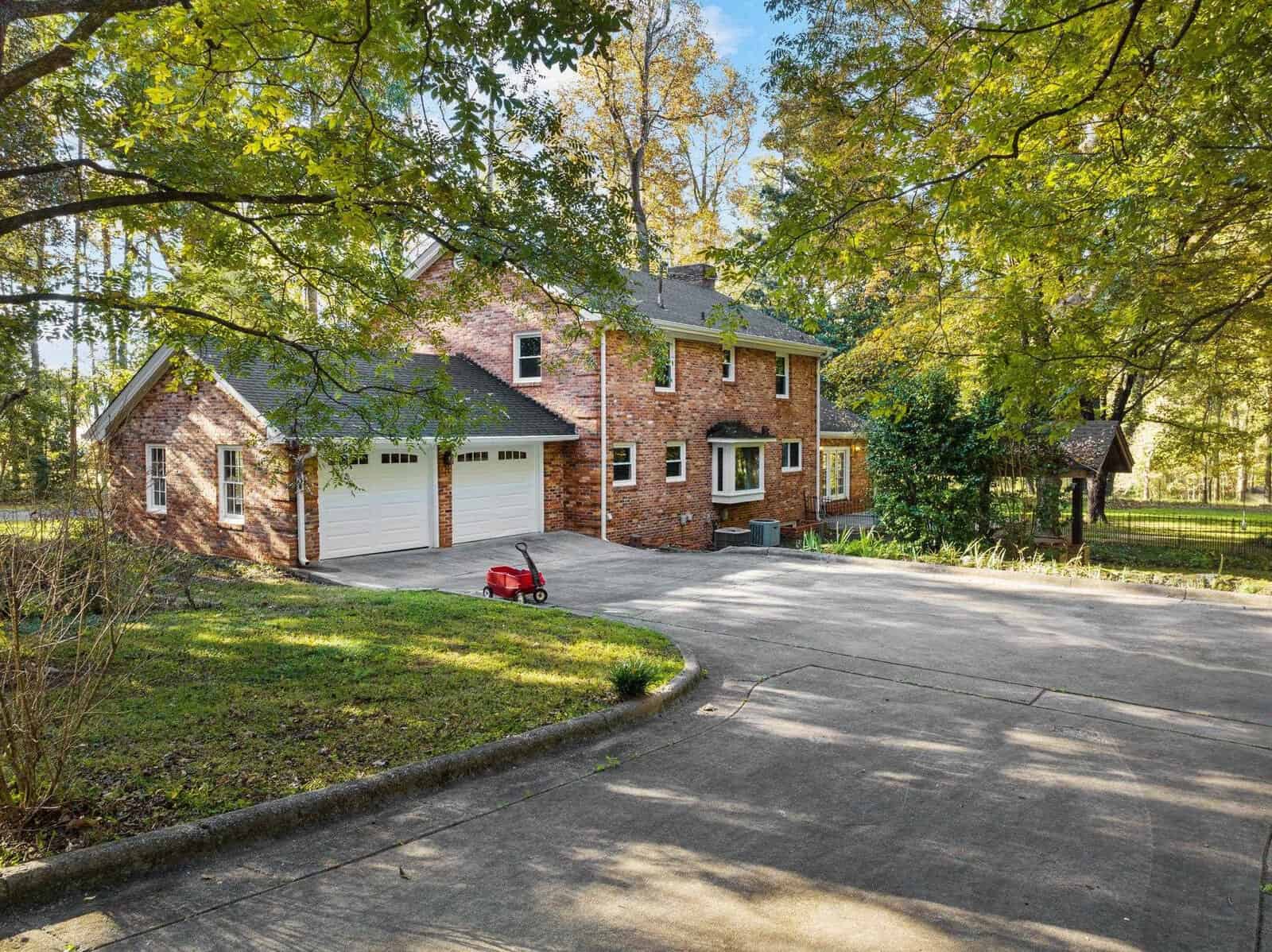 A two-story brick house with a large driveway surrounded by trees. A red toy car is parked near the house. The setting is sunny with shadows of leaves on the driveway, and the yard is bordered by a fence.