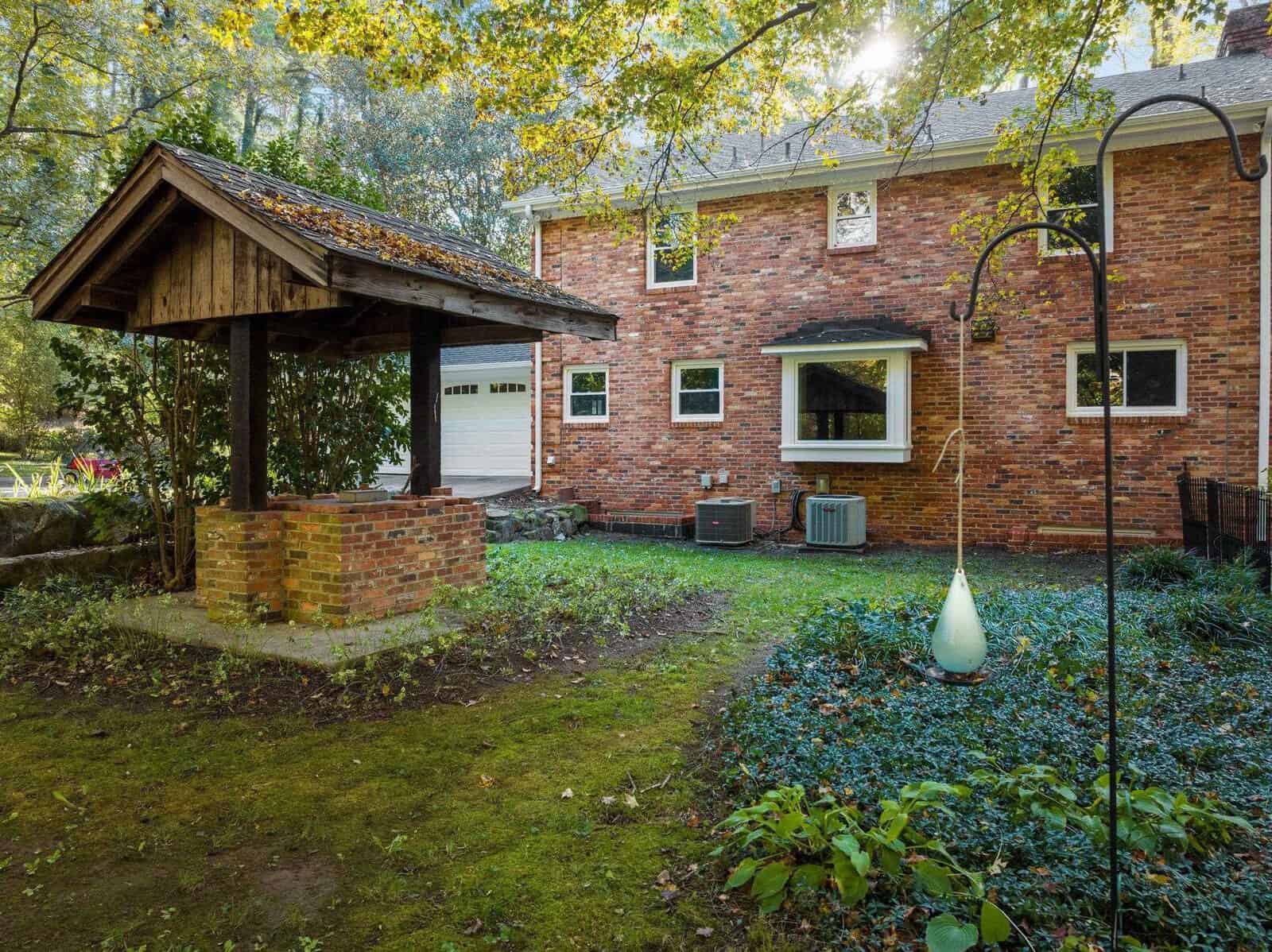 Brick house with a two-car garage, a rustic well with a pitched roof in the backyard, surrounded by trees and greenery. A hanging bird feeder is in the foreground on a grassy lawn. Sunlight filters through the leaves.