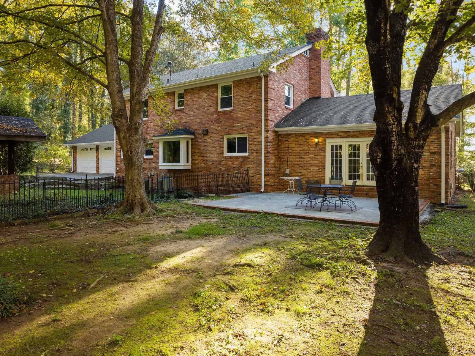 A two-story brick house with large windows has a spacious backyard. The outdoor area features a patio with patio furniture and is surrounded by trees, casting dappled sunlight on the grass. A garage is visible on the left side.