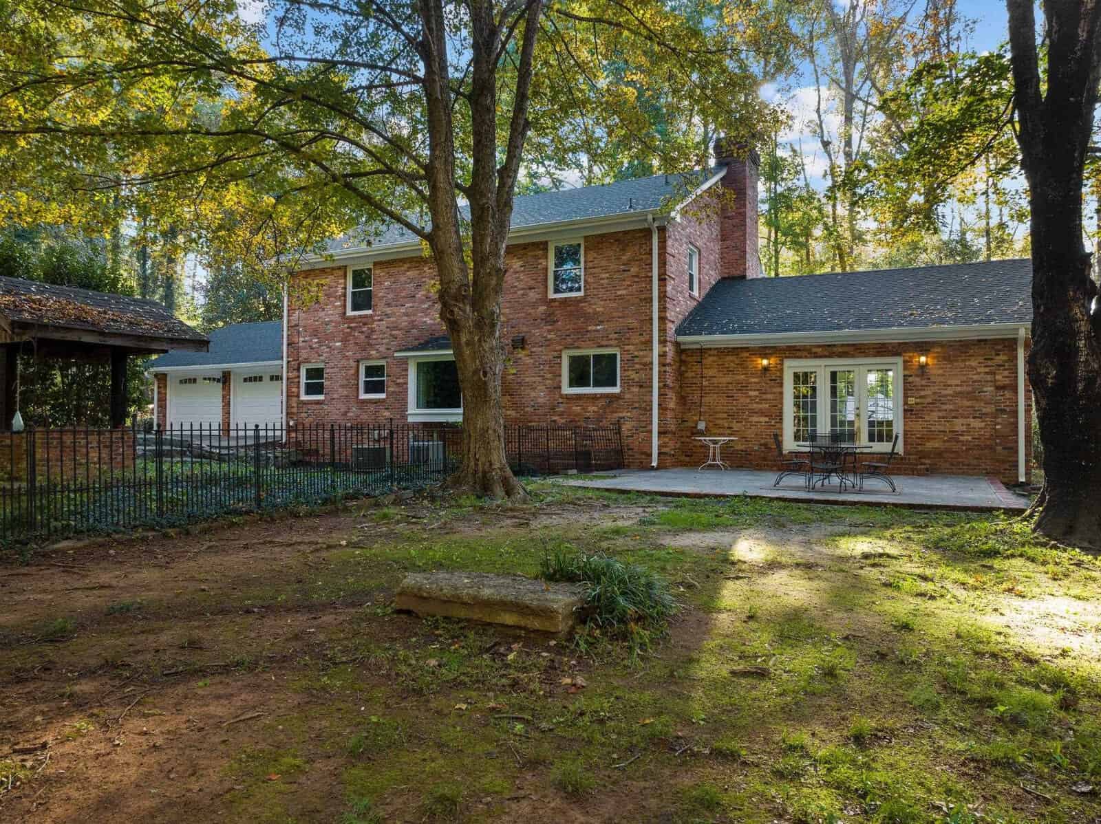 Two-story brick house surrounded by trees, featuring a fenced yard and a spacious patio with outdoor furniture. A large tree and grassy area are in the foreground, creating a peaceful backyard setting.