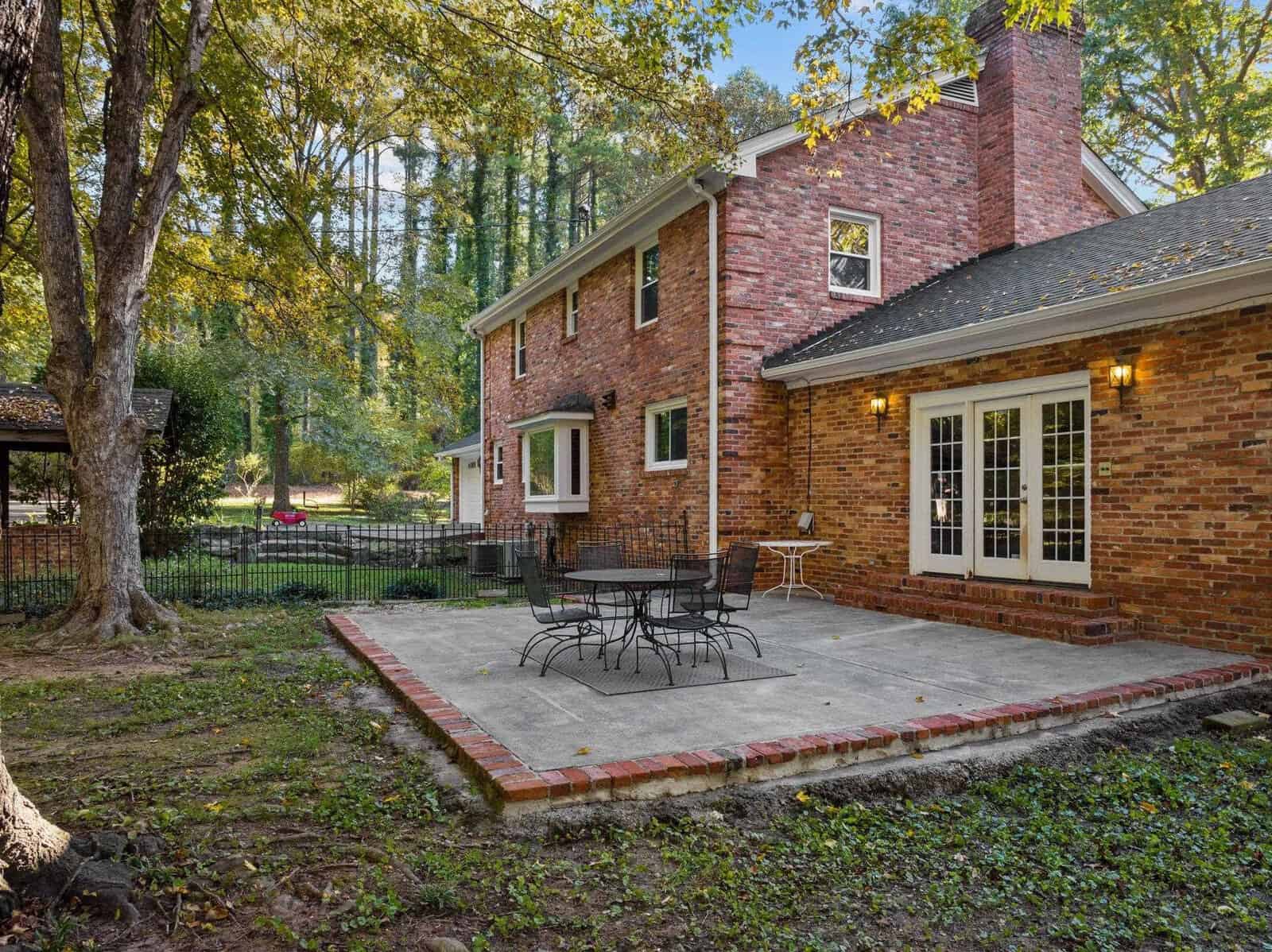 A brick house with a spacious backyard features a large patio area with outdoor furniture, surrounded by tall trees and a grassy lawn. The home has multiple windows and a chimney, creating a cozy, inviting atmosphere.