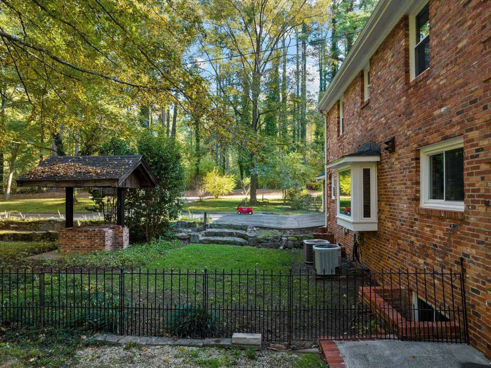 A brick house with a fenced yard features a grassy area and a small covered well. Tall trees surround the property, and a red toy car sits in the background near a stone pathway. The scene is peaceful and sunlit.