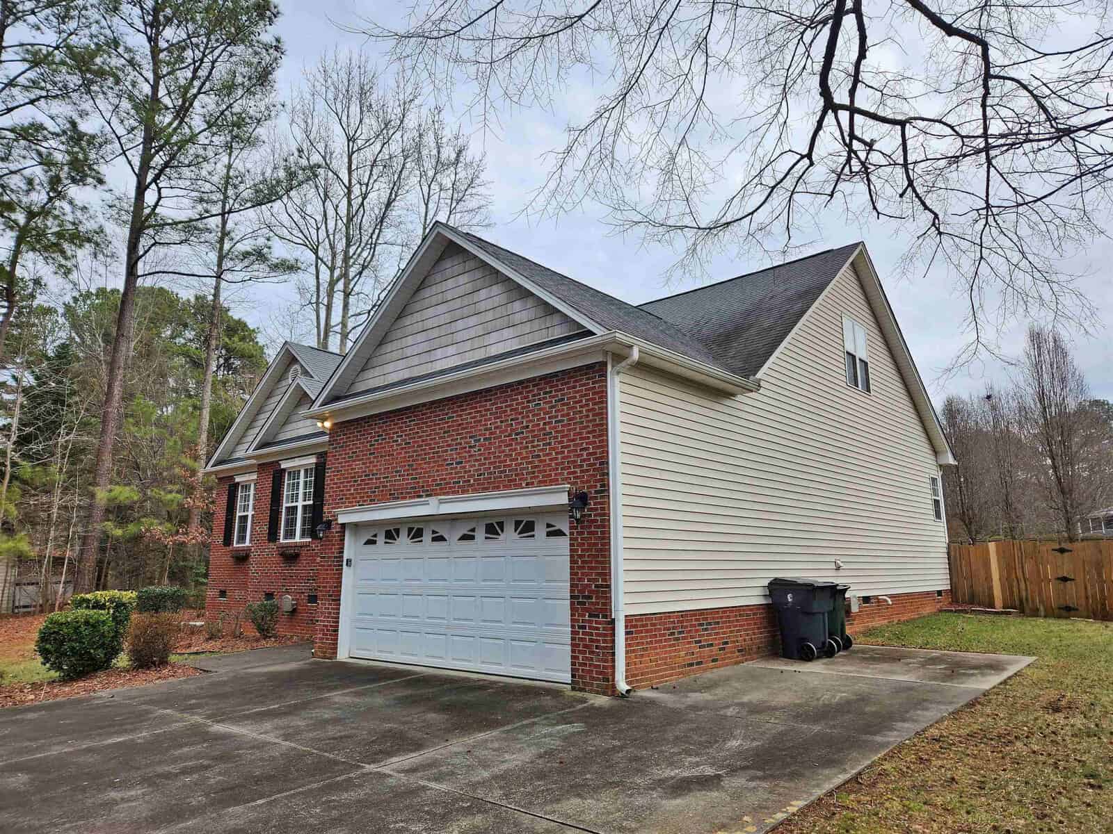A suburban house with red brick and beige siding, featuring a two-car garage with a white door. The driveway is concrete, and there are two black trash bins. The property is surrounded by trees and a wooden fence.