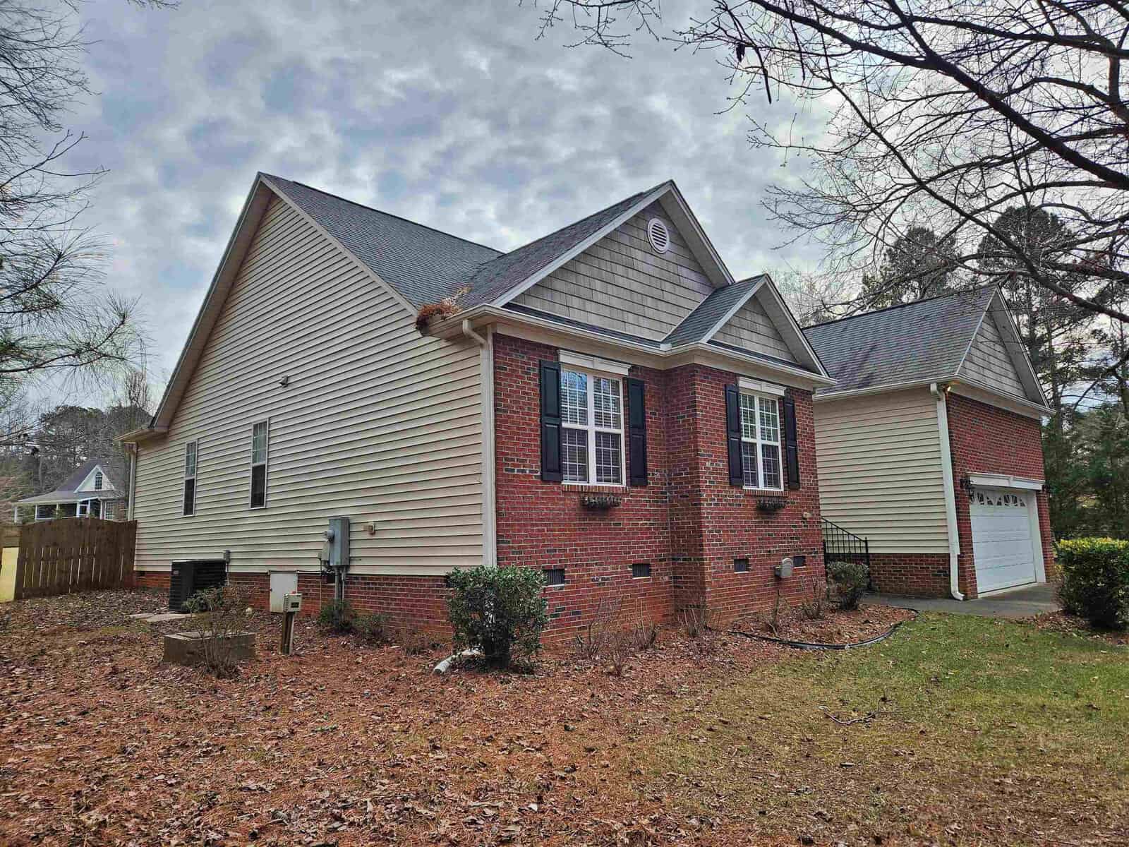 A single-story house featuring red brick and beige siding. It has white-framed windows and a two-car garage. The roof is gray, and the house is surrounded by a lawn with scattered trees and dry leaves.
