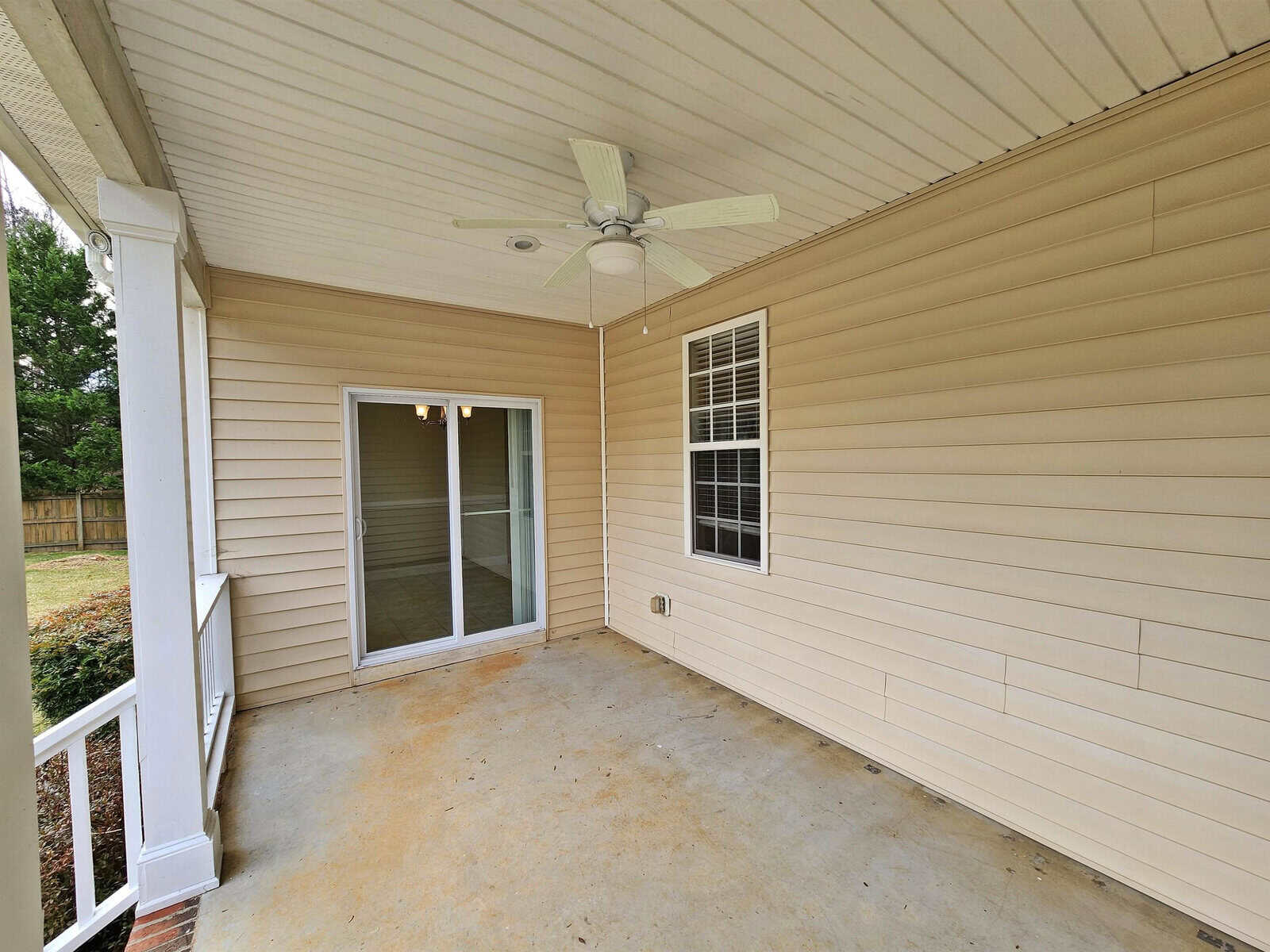A covered patio with beige siding features a ceiling fan and a sliding glass door leading inside. The floor is concrete, and there's a white-framed window on the wall. The area overlooks a grassy backyard with a wooden fence.