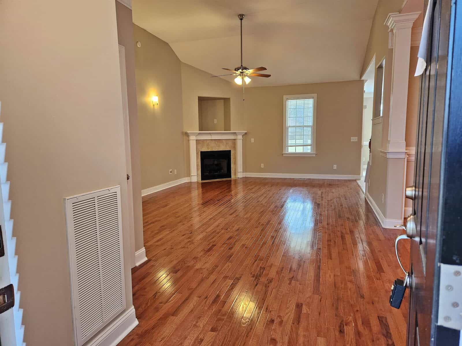 A spacious living room with polished wooden floors and beige walls. It features a ceiling fan, a large window, and a fireplace framed by a small alcove. A decorative light fixture is on the wall, and an air vent is visible near the entrance.