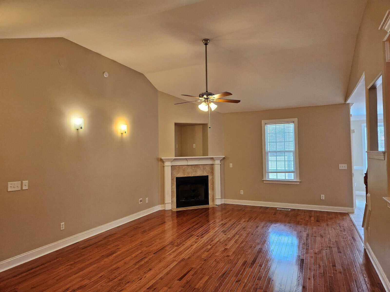 A spacious living room with wooden floors, a high ceiling, and beige walls. The room features a ceiling fan, two wall lights, a fireplace with a mantle, and a window allowing natural light.