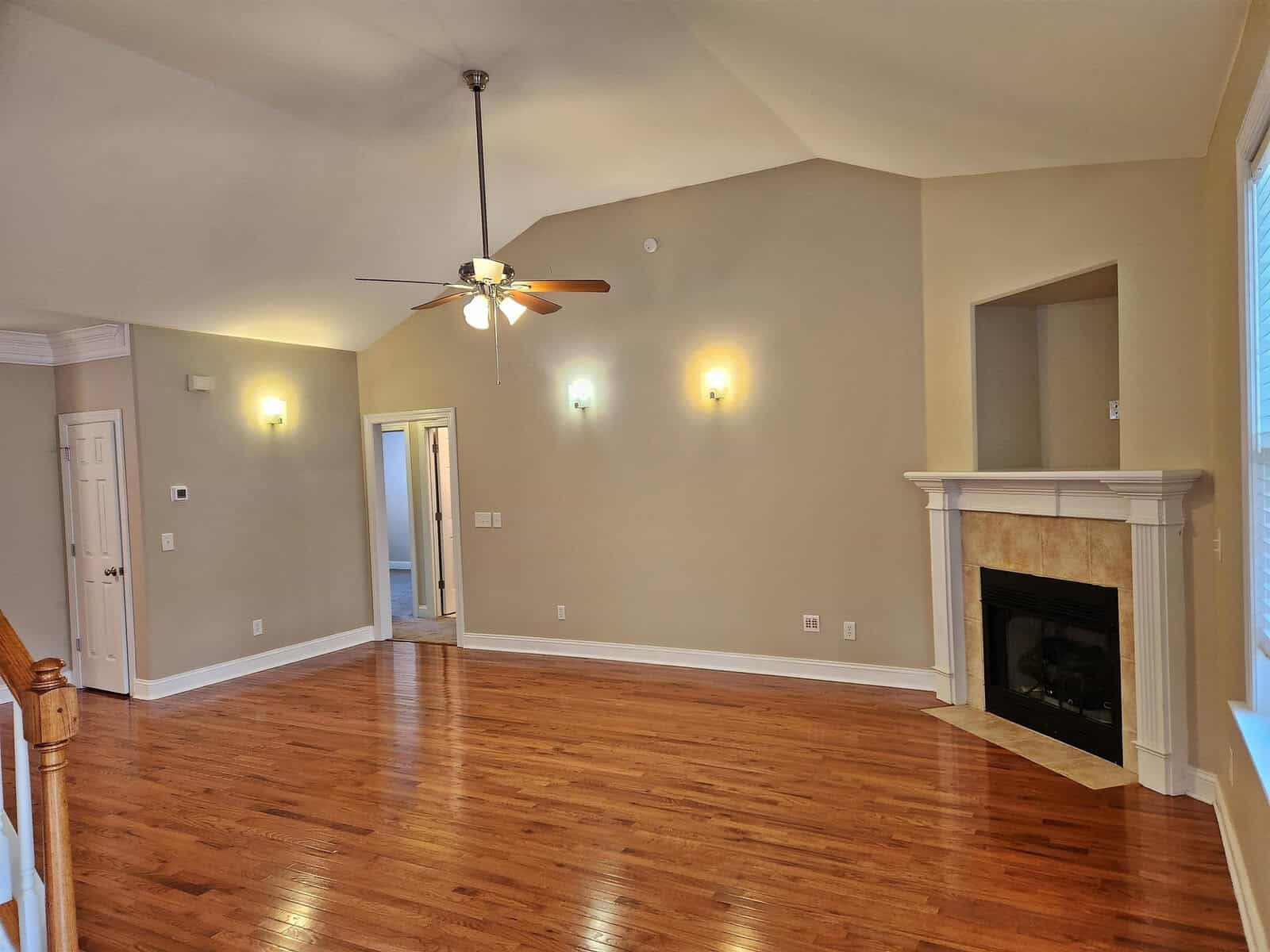 Spacious living room with hardwood floors, beige walls, and a vaulted ceiling. Features a ceiling fan with lights, a white mantle fireplace on the right, and a doorway leading to another room. Natural light enters through a window on the right.