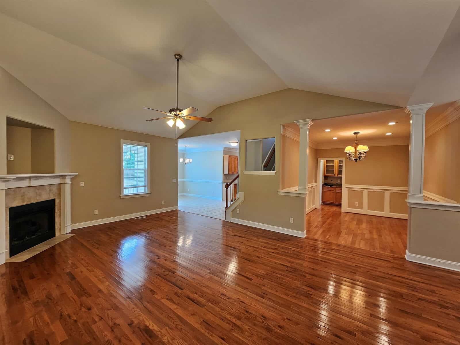 Spacious living area with vaulted ceiling, ceiling fan, and wooden floors. Features a fireplace and opens into a bright, tiled dining area with a chandelier. Neutral walls and large windows enhance natural light.