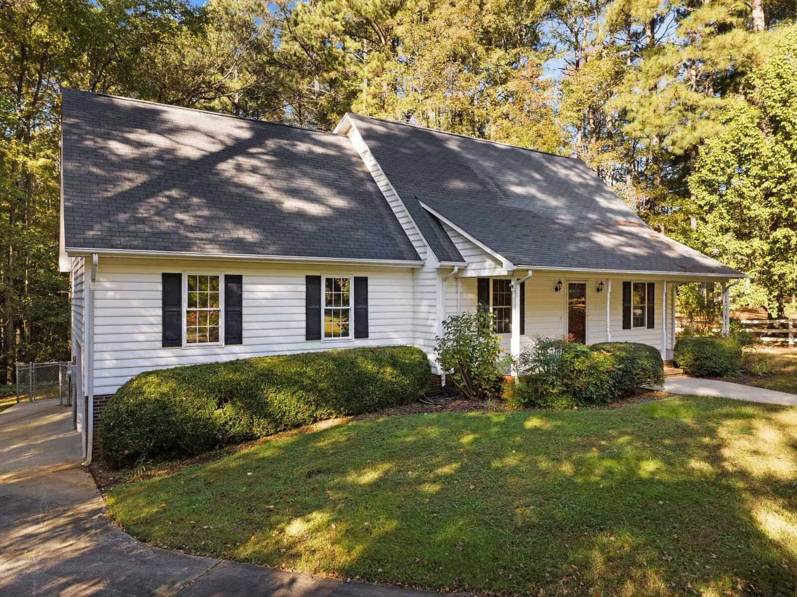 Nestled in a wooded area under a clear sky, this charming white house on 4126 Samuels Court boasts a dark gray gabled roof and black shutters. Surrounded by lush green bushes and trees, it features a welcoming front porch with white railings and a curved driveway typical of Oxford elegance.
