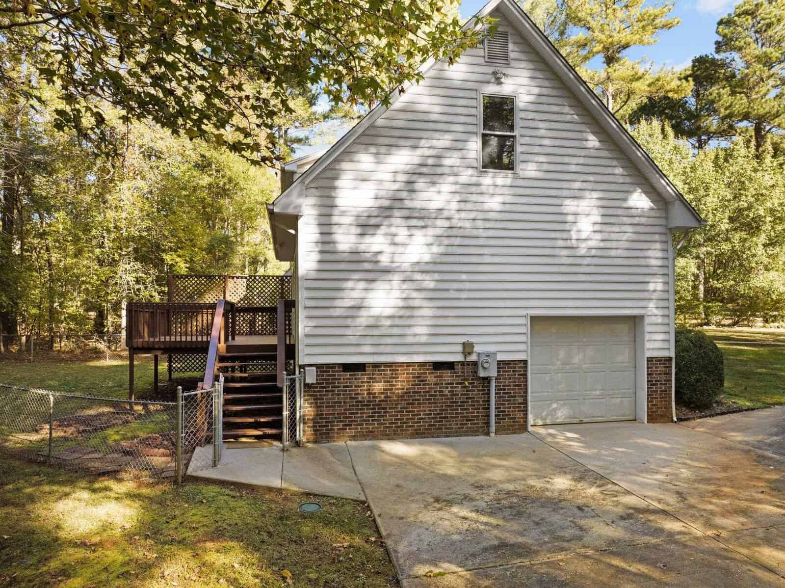 A house with white siding and a brick foundation is shown. There's a garage on the lower level, a wooden deck on the side, and it is surrounded by trees and a driveway. A chain-link fence is visible near the deck.