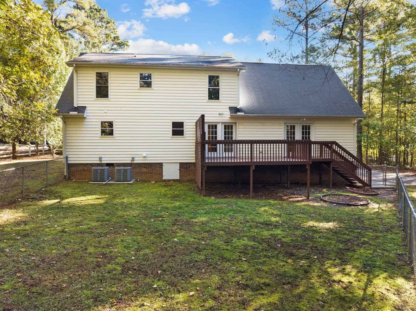 A two-story, light-colored house with a wooden deck, surrounded by trees and a fenced yard. The roof is gray, and the area is grassy with patches of sunlight filtering through the trees. The day is clear and sunny.