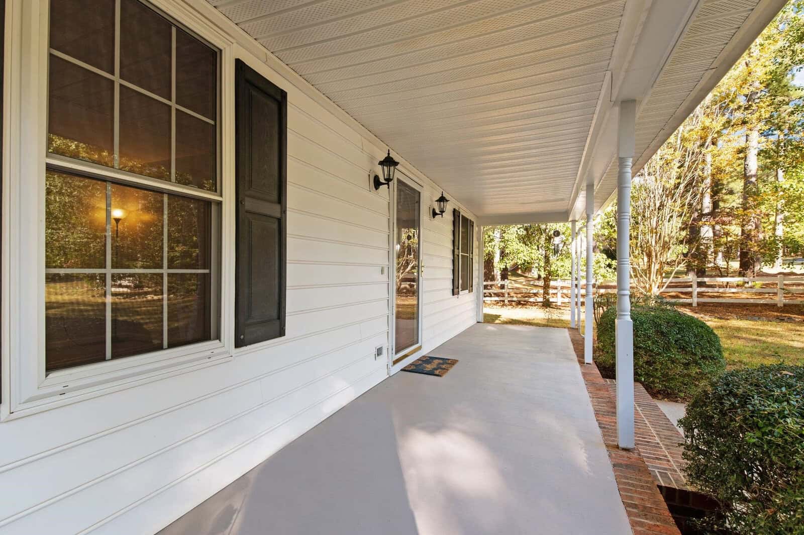A white house porch with a covered ceiling is lined with black framed windows and lantern-style lights. The porch floor is concrete, leading to a view of a green lawn and trees bordered by a wooden fence.