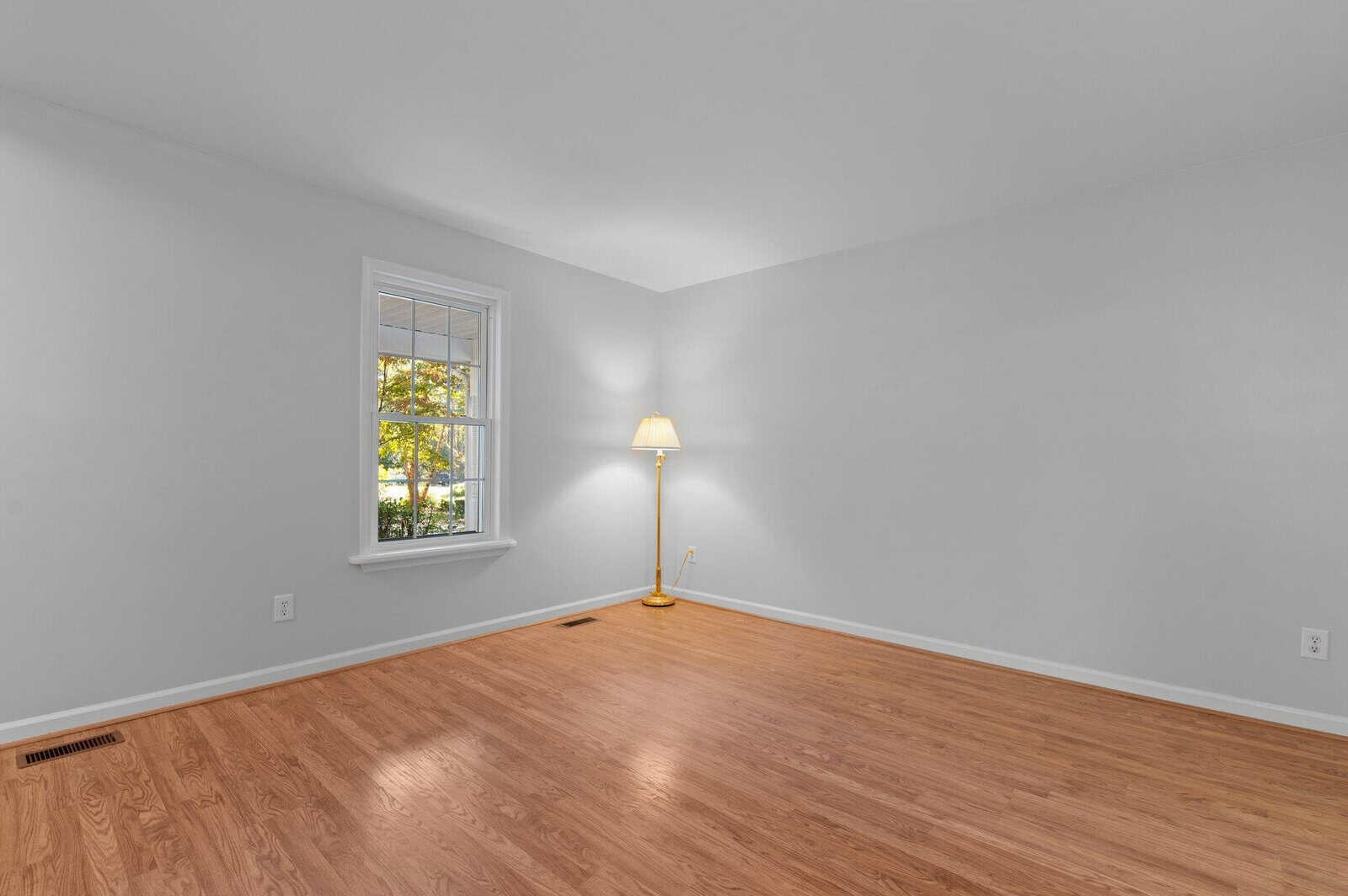 A minimalistic room with light gray walls and wooden flooring. A single window on the left lets in natural light. A floor lamp with a beige shade is situated in the corner, casting a warm glow. The space is empty and spacious.