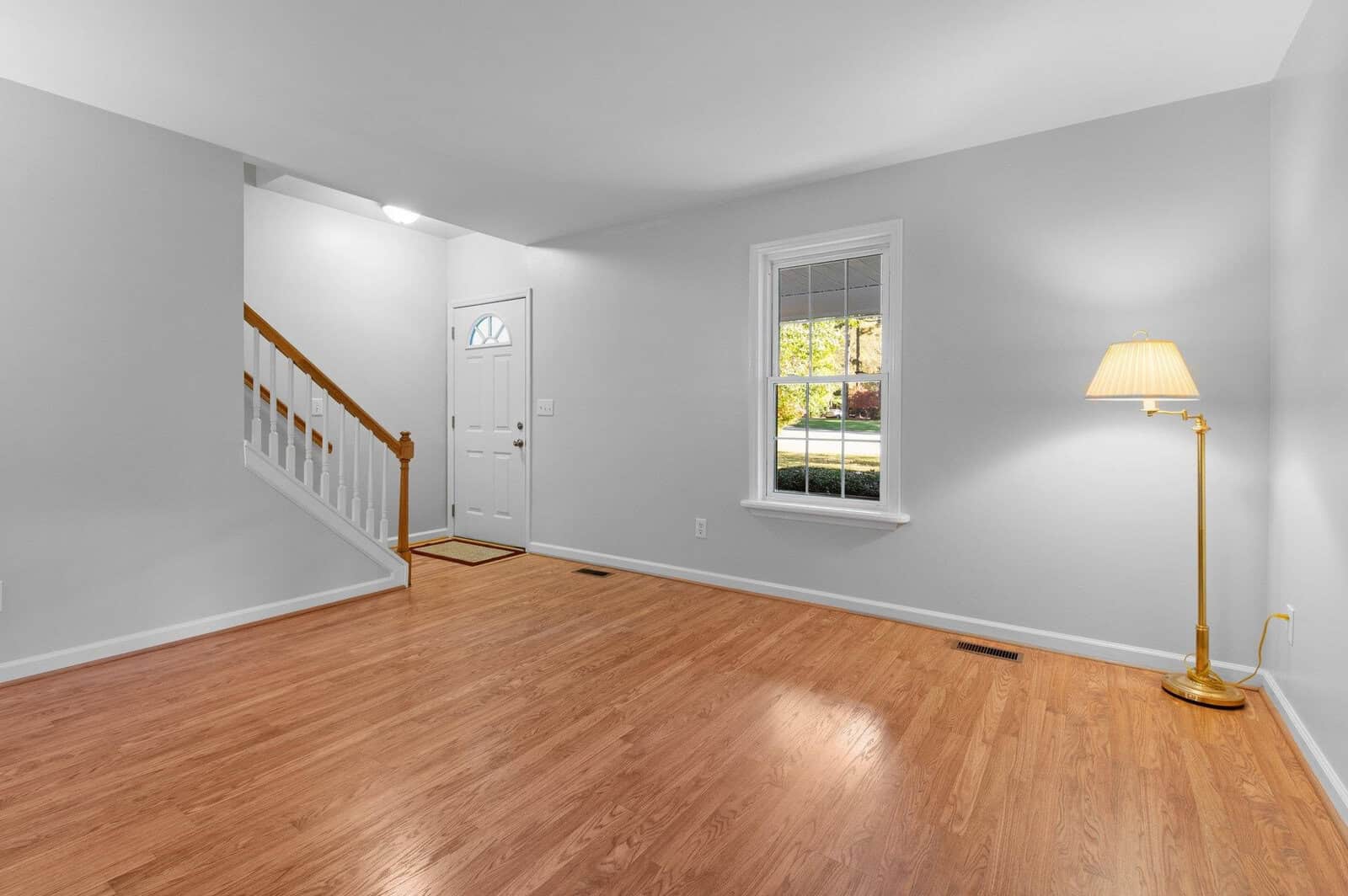 A bright, empty living room with light gray walls and hardwood flooring. There is a wooden staircase to the left, a large window, and a gold floor lamp on the right. The entrance door is visible near the stairs.