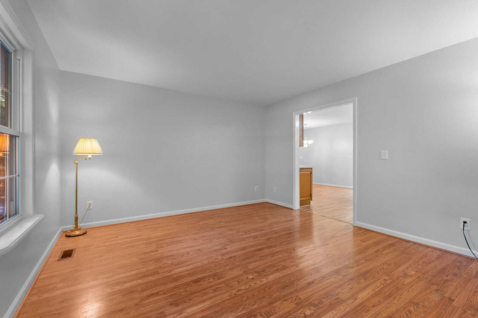 Empty room with light gray walls and hardwood floors, featuring a window on the left and a golden floor lamp. A doorway offers a view into an adjacent room with a hanging light fixture and visible kitchen cabinetry.