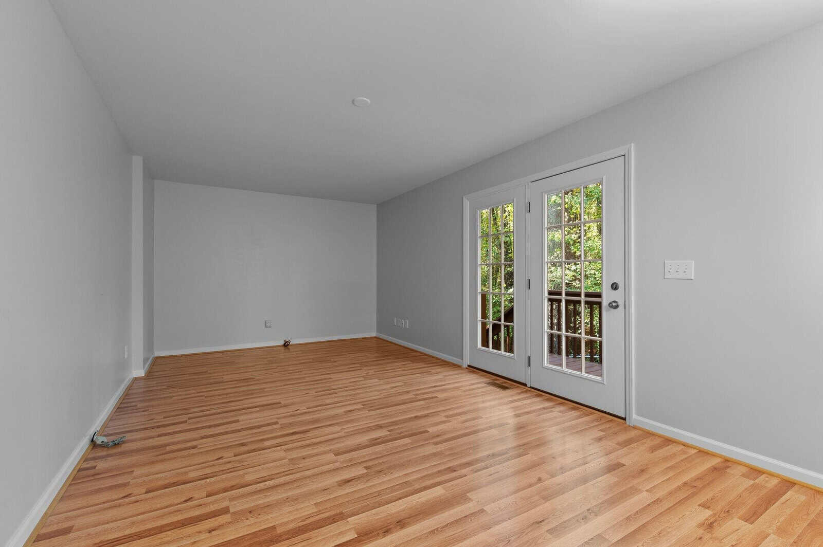 An empty room with light wood flooring, gray walls, and French doors leading to a small balcony. Natural light is coming through the doors, illuminating the space.