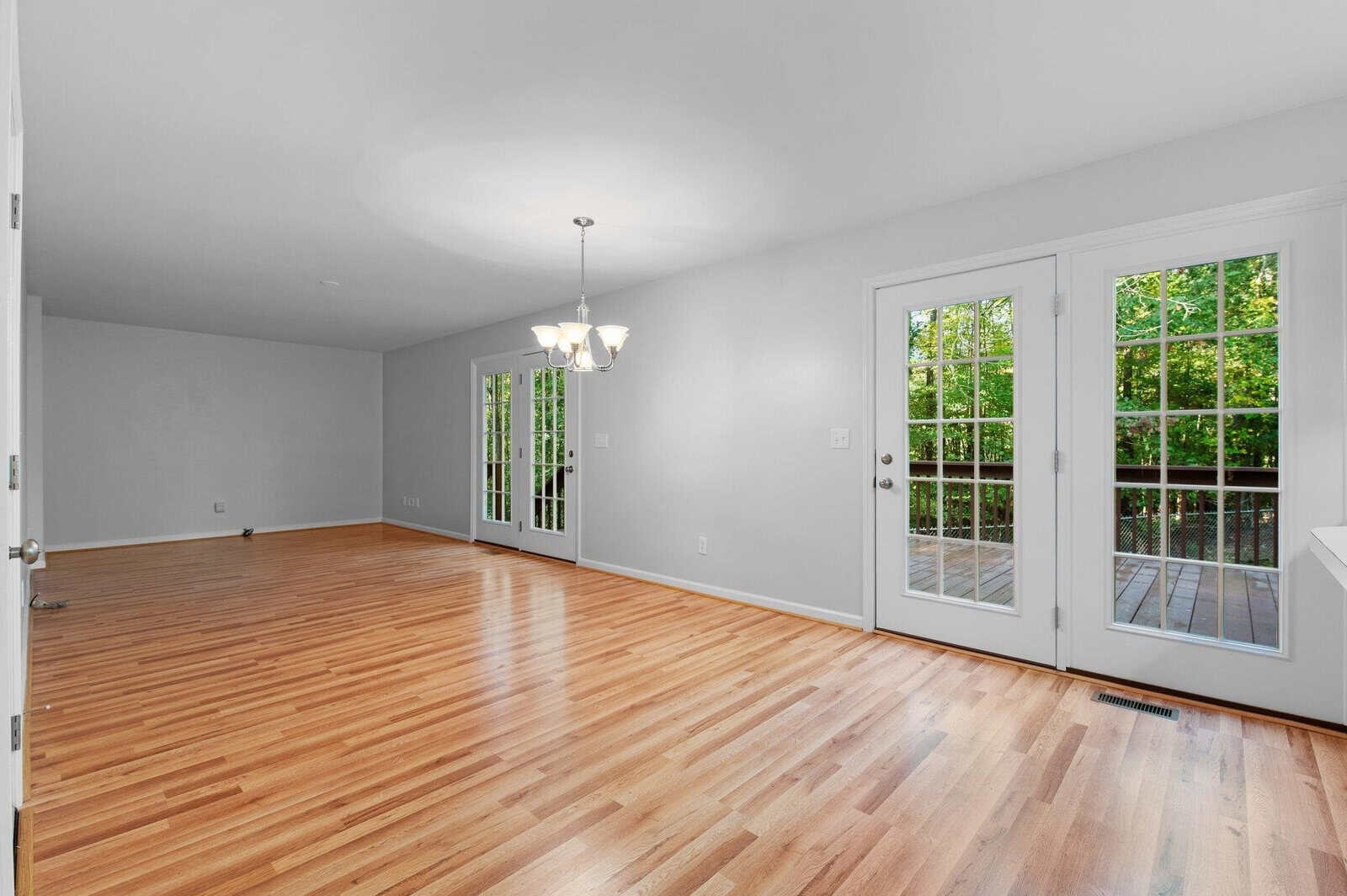 A bright, empty room with wooden flooring, white walls, and a modern chandelier. French doors with glass panes open to a deck, offering a view of green foliage outside. Natural light pours in through the doors.