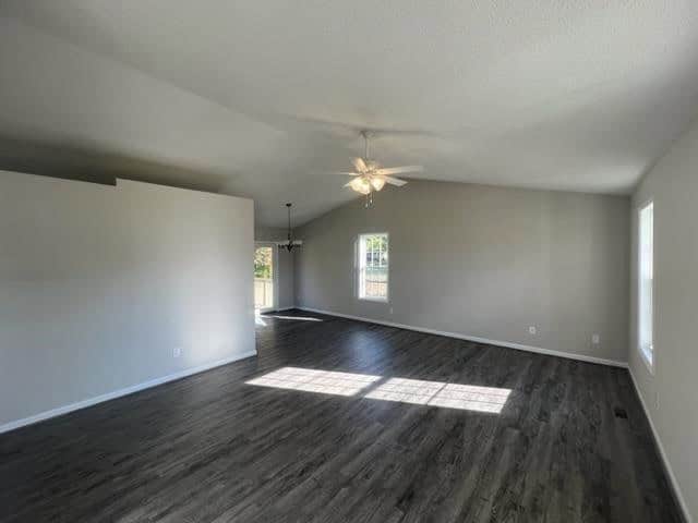 Empty living room with gray walls, dark laminate flooring, and a ceiling fan with lights, illuminated by natural light from windows.