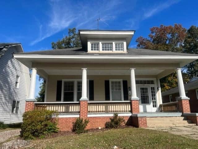 A white single-story house with a broad porch supported by four columns under a large gabled roof, set against a clear sky. the house has multiple windows and a grassy front yard.