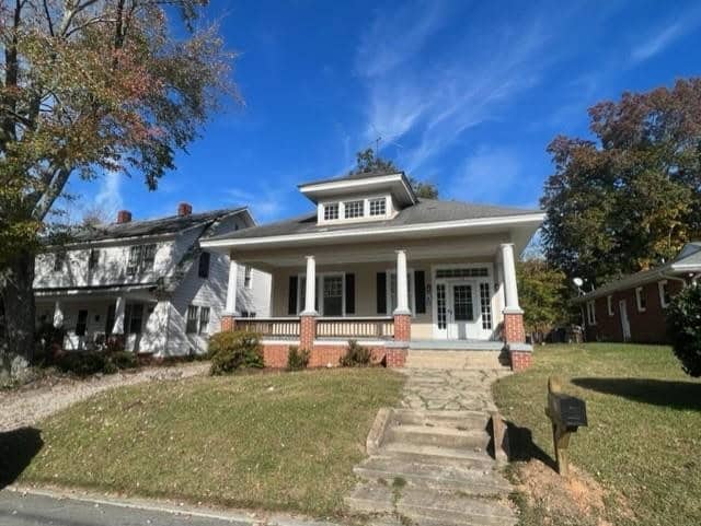 A white colonial house with a large front porch, red brick steps, and a balustrade, surrounded by mature trees under a clear blue sky.