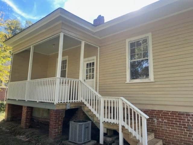 A single-story house with beige siding, featuring a covered front porch with white railings and a staircase leading up to it. there's an air conditioning unit near the base of the porch.