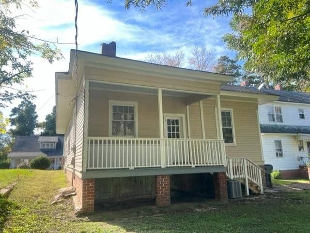 A quaint yellow house with white trim, featuring a covered front porch with balustrades. the house is partially elevated on a brick foundation with a visible crawl space. there are trees around and a neighboring house visible.