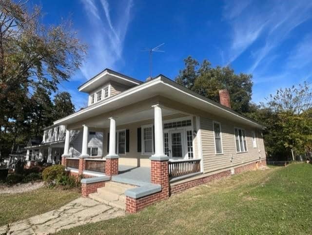 A single-story cream-colored house with a wrap-around porch, white columns, and a red brick chimney under a clear blue sky. there are trees and a lawn in the foreground.